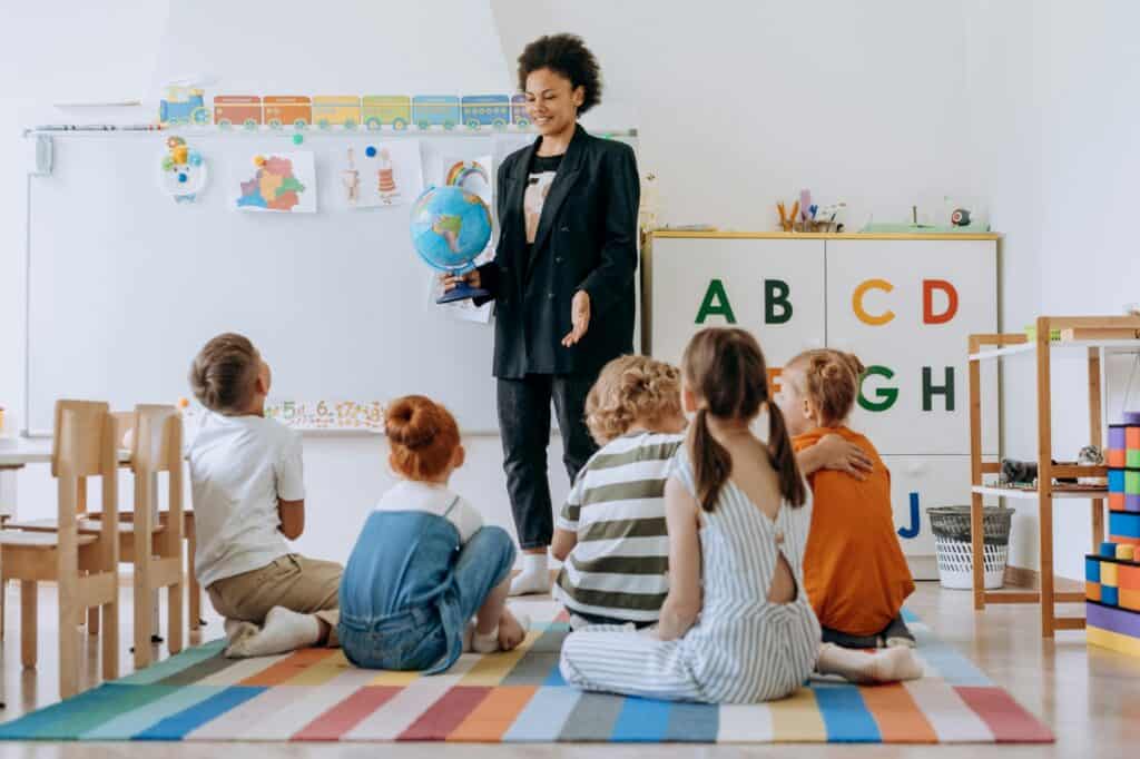 The teacher is in front of the class while the young students are sitting on the floor in the concept of 'top schools in Wood Green'.